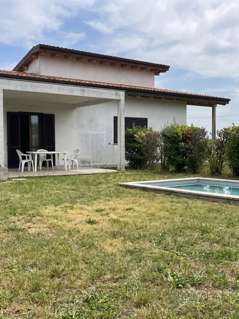 Simulazione con AI della villa prima del progetto, con prato incolto, sedie di plastica sotto il portico e piscina senza arredi